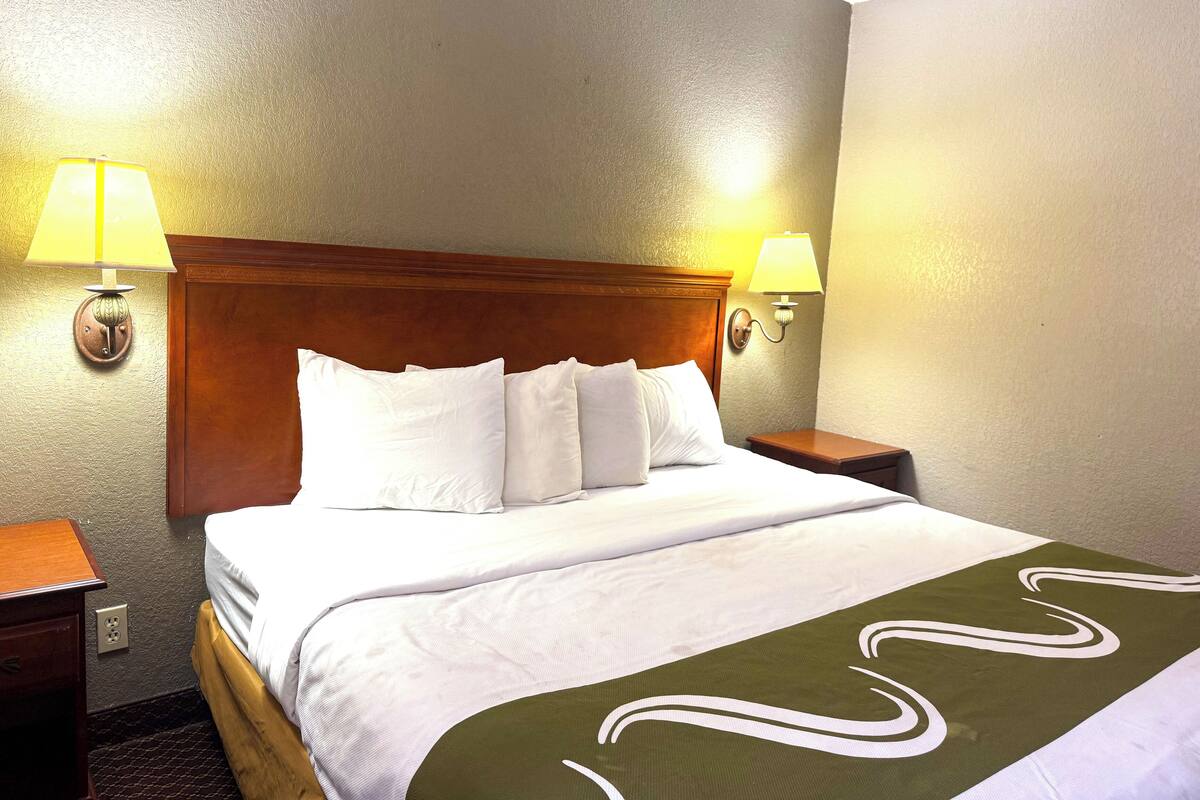 Una cama bien tendida con sábanas blancas y una manta verde, dos lámparas de noche y un cabecero de madera.
