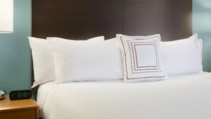 Un lit bien fait, avec du linge de lit blanc et un coussin à motifs géométriques, une tête de lit en bois et une horloge numérique affichant l’heure.