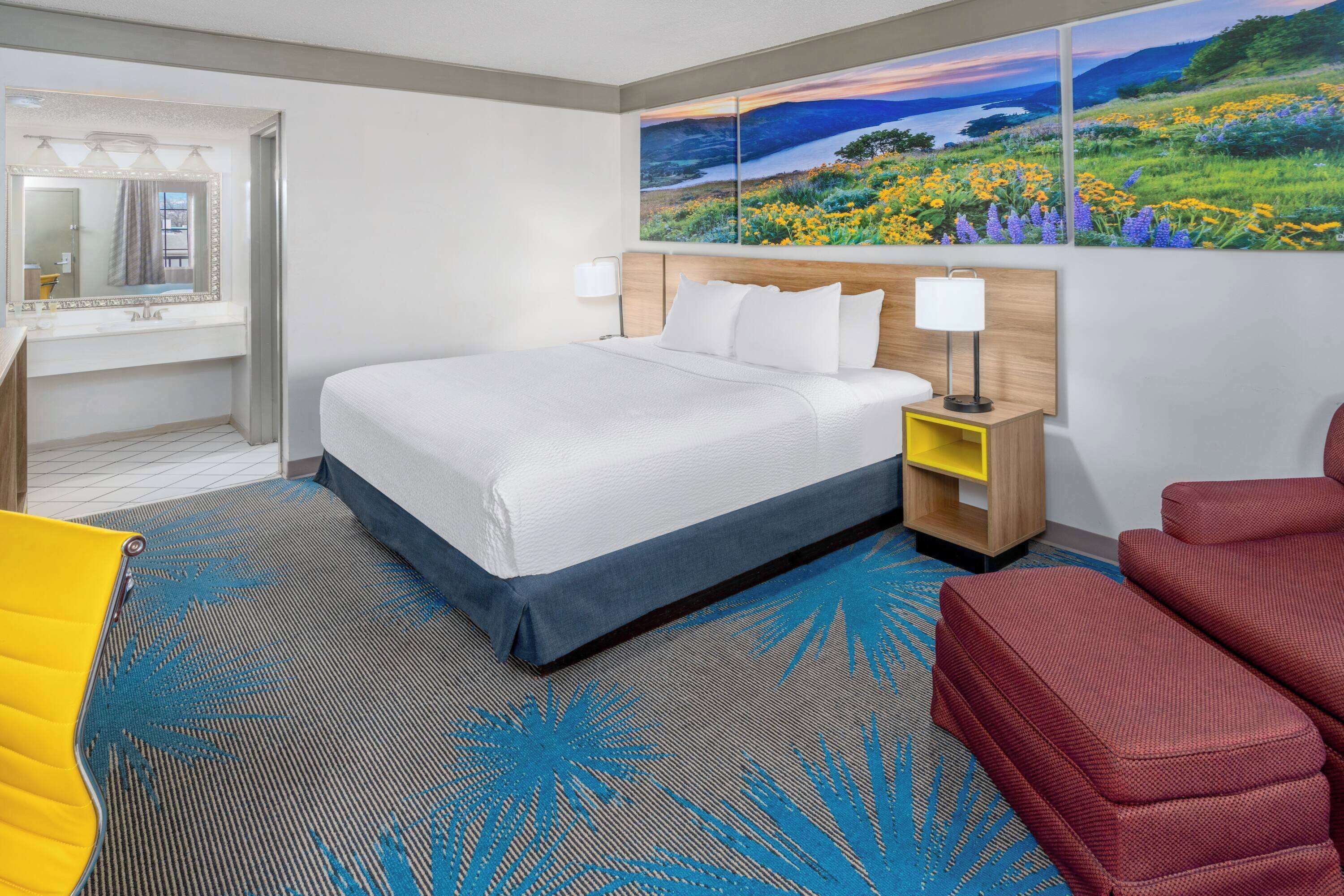 A hotel room with a bed, a desk with a mirror, a nightstand, a red sofa, and a large landscape painting on the wall.