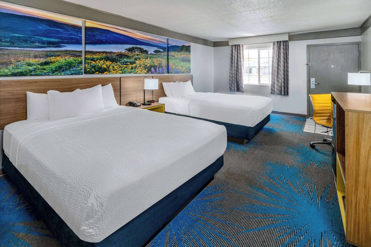 A hotel room with two beds, a desk, a chair, and a large window with a view of a landscape painting.