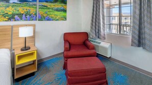 Habitación de hotel con una silla roja, una mesita de noche de madera con una lámpara, un ventanal con cortinas y un cuadro de un campo.