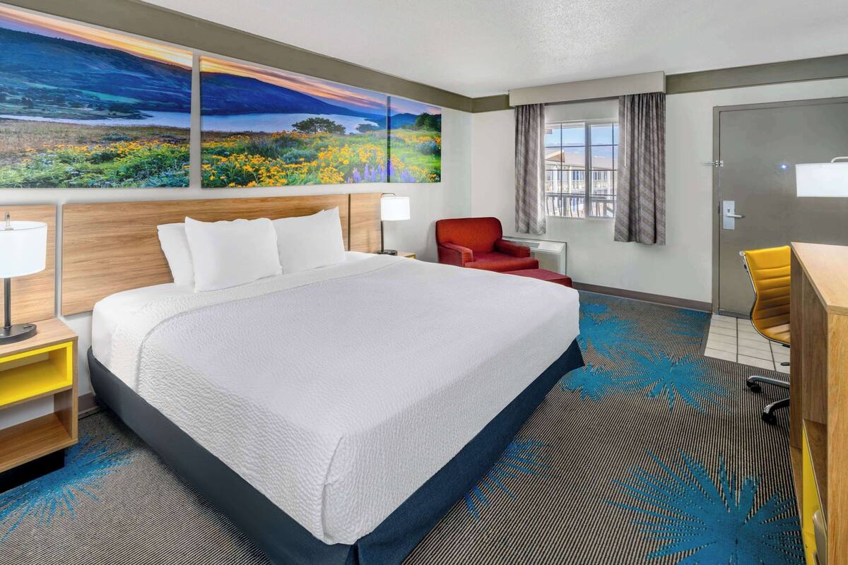A hotel room with a large bed, a desk, a chair, a red armchair, and a painting of a landscape on the wall.