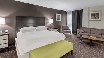 Habitación de hotel con una cama grande, un sofá, una silla, un escritorio con una lámpara y un cuadro en la pared.