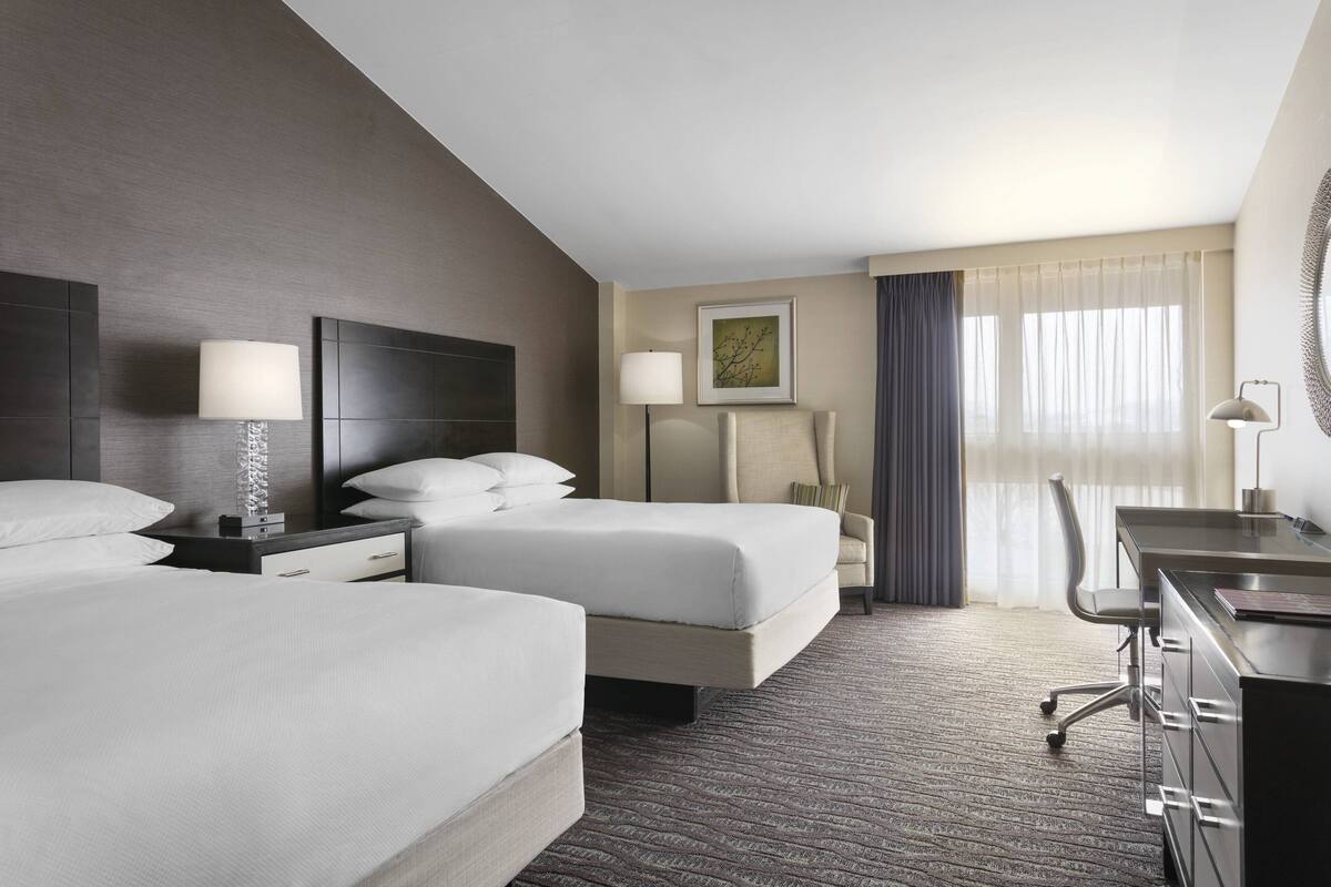 A hotel room with two beds, a desk, a chair, and a large window with curtains.