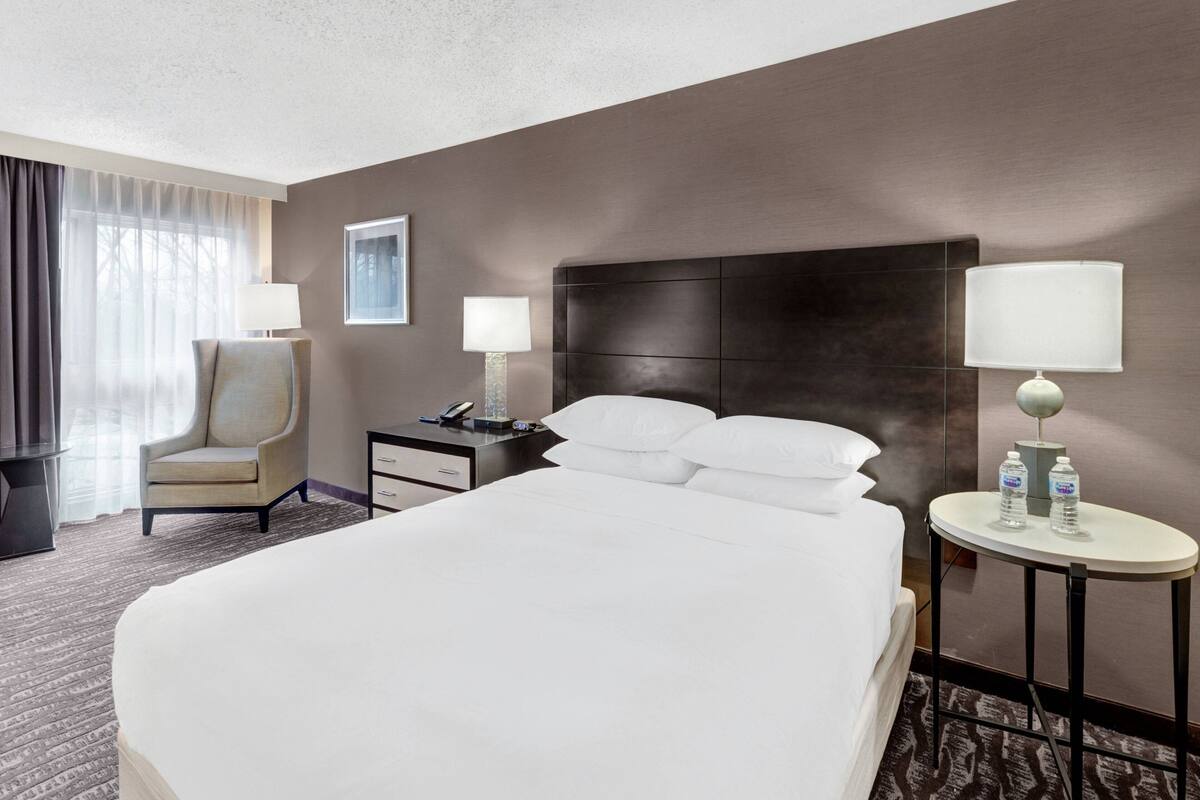 A hotel room with a large bed, a chair, a nightstand with a lamp, and a small table with water bottles.