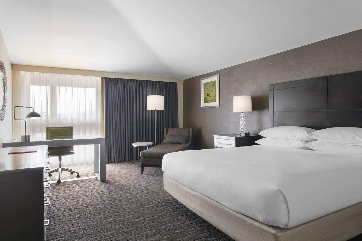 A hotel room with a bed, a desk, a chair, a nightstand, and a lamp.