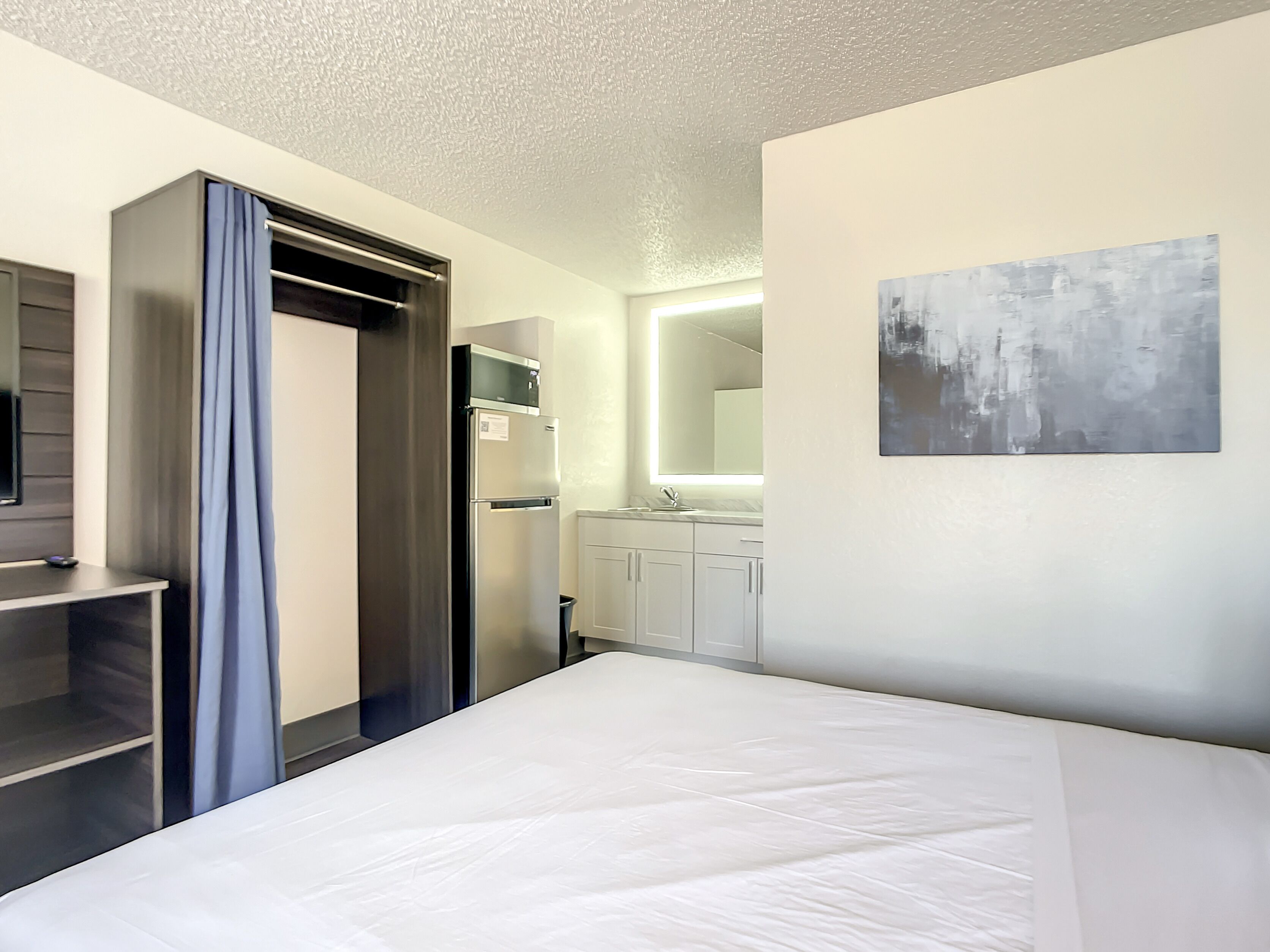A hotel room with a bed, a built-in desk, a refrigerator, a mirror, and a painting on the wall.