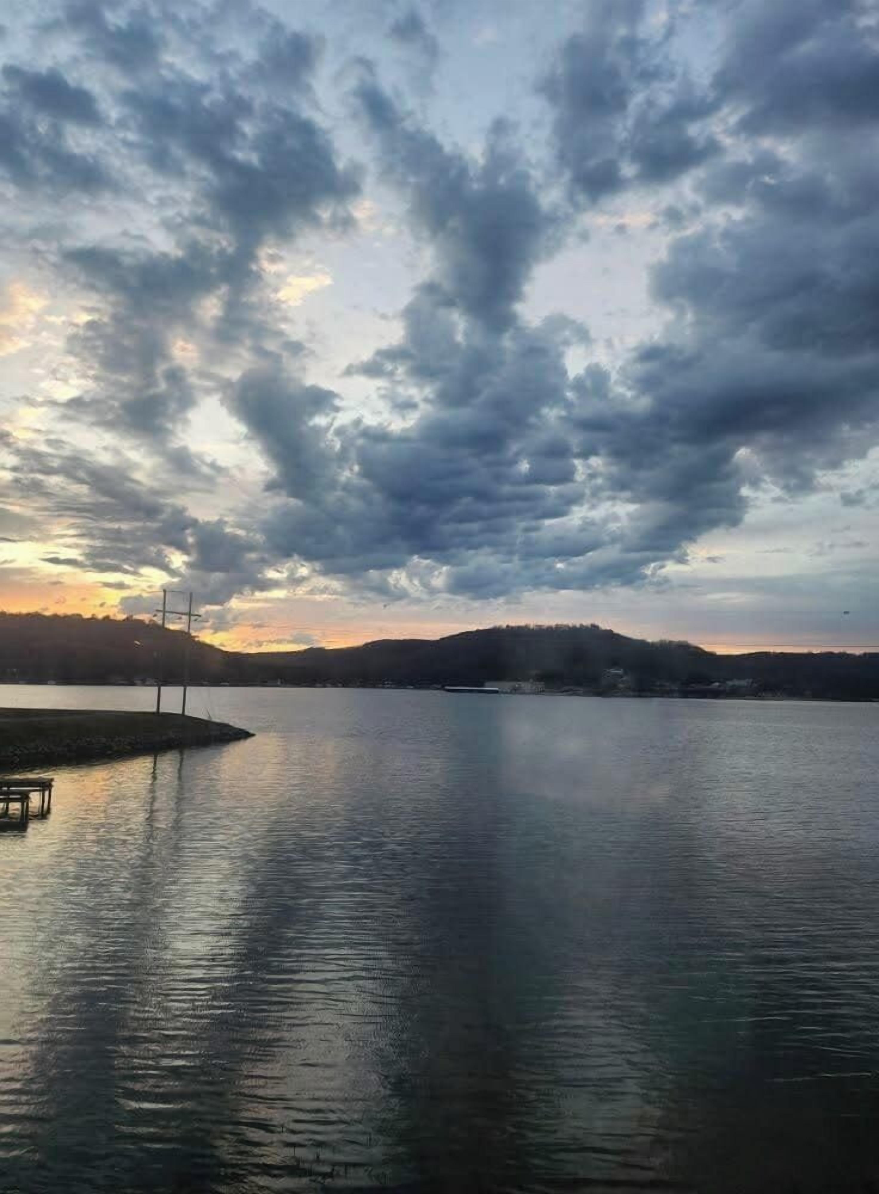 A calm lake with a dock and a bench, surrounded by hills and a cloudy sky.