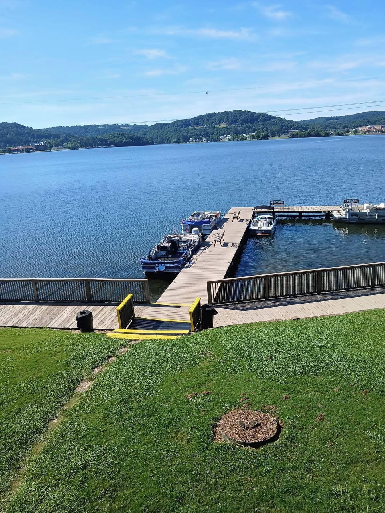 Un muelle con barcos, un área verde y un lago con colinas al fondo.