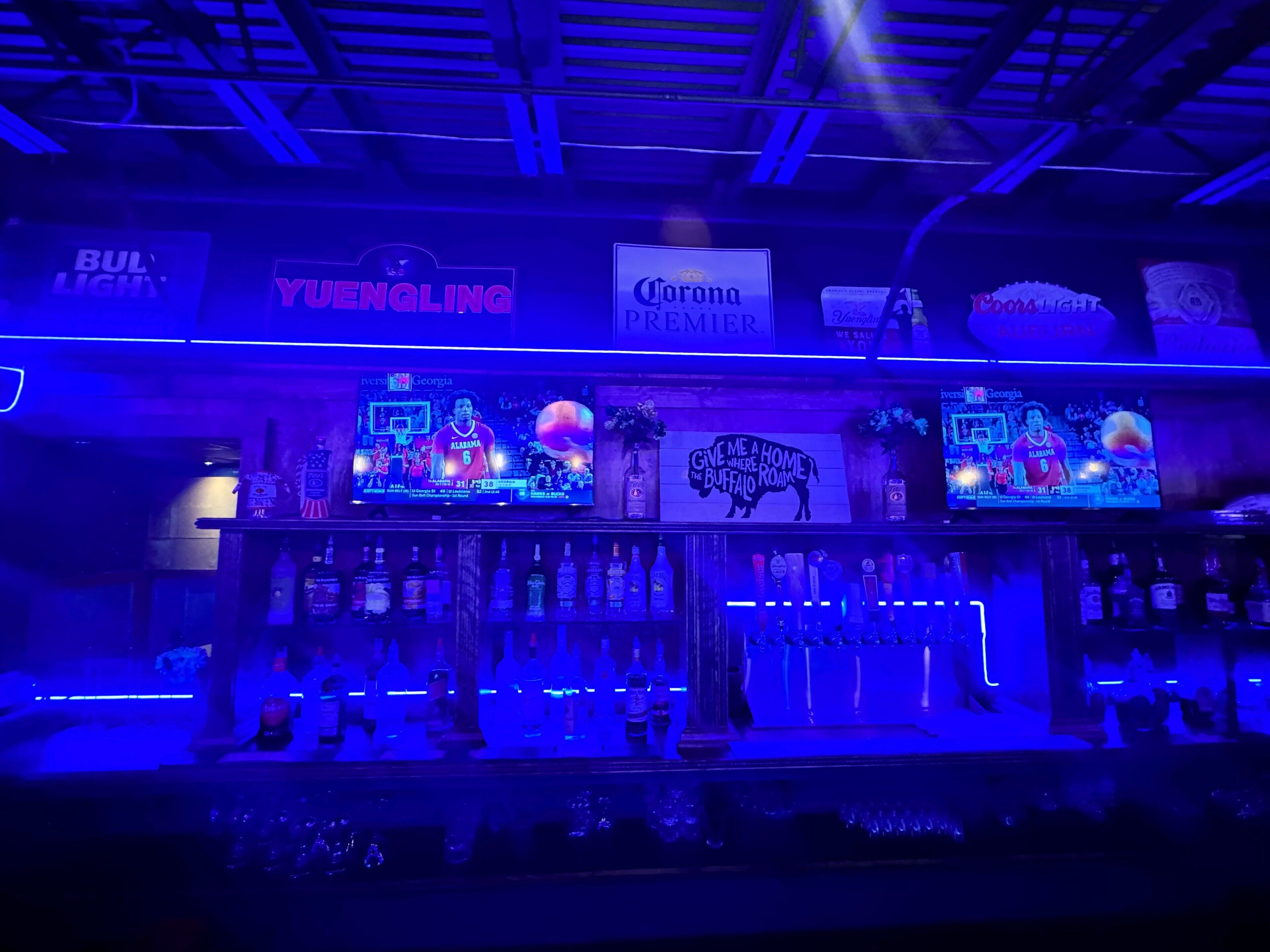 A bar area with a large screen displaying sports content, shelves stocked with various liquor bottles, and branded signs for 'Yuenling' and 'Corona Premier'.