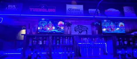 A bar area with a large screen displaying sports content, shelves stocked with various liquor bottles, and branded signs for 'Yuenling' and 'Corona Premier'.