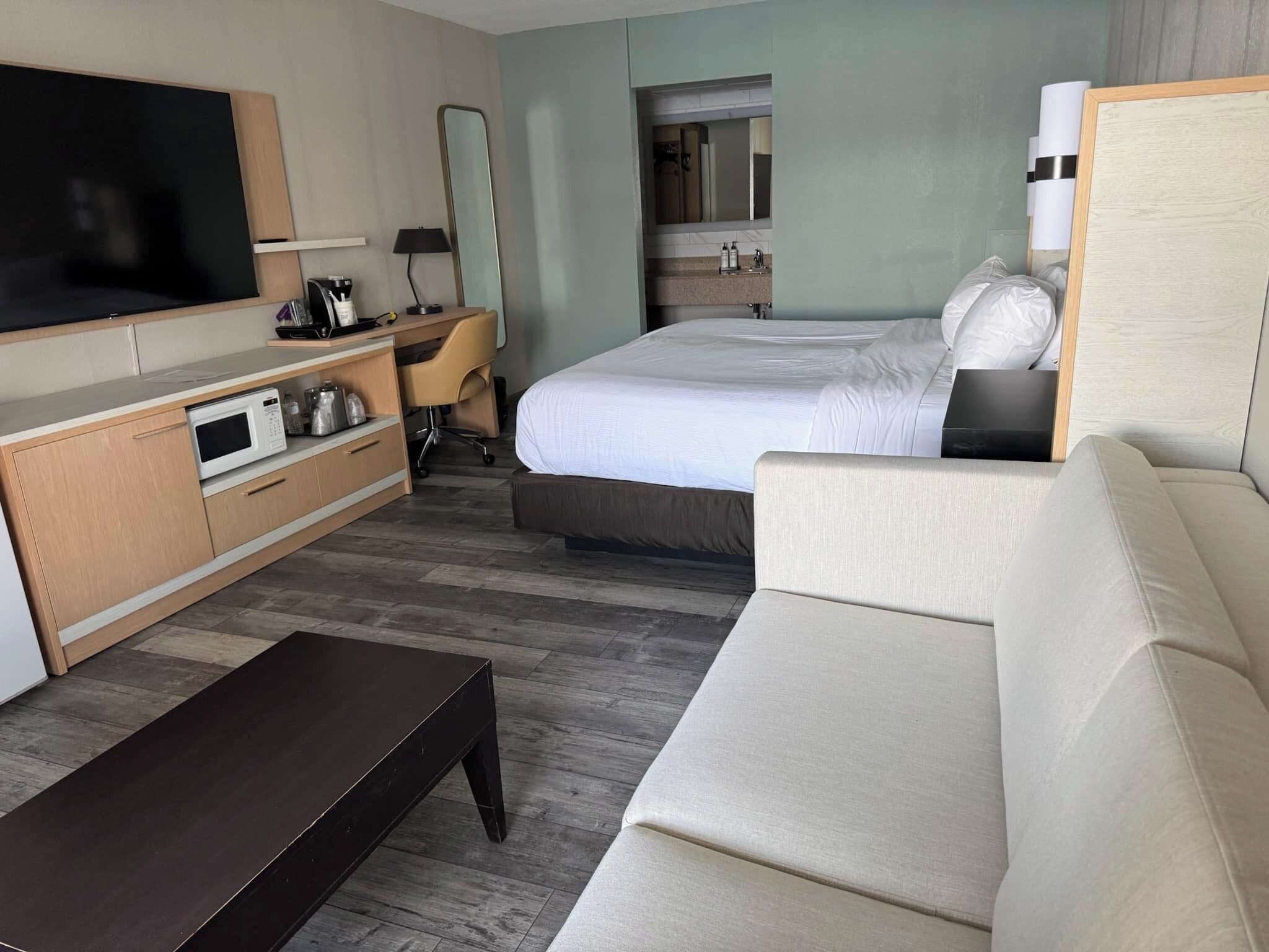 A modern hotel room with a large bed, a sofa, a TV, a desk, and a bathroom visible through a door.