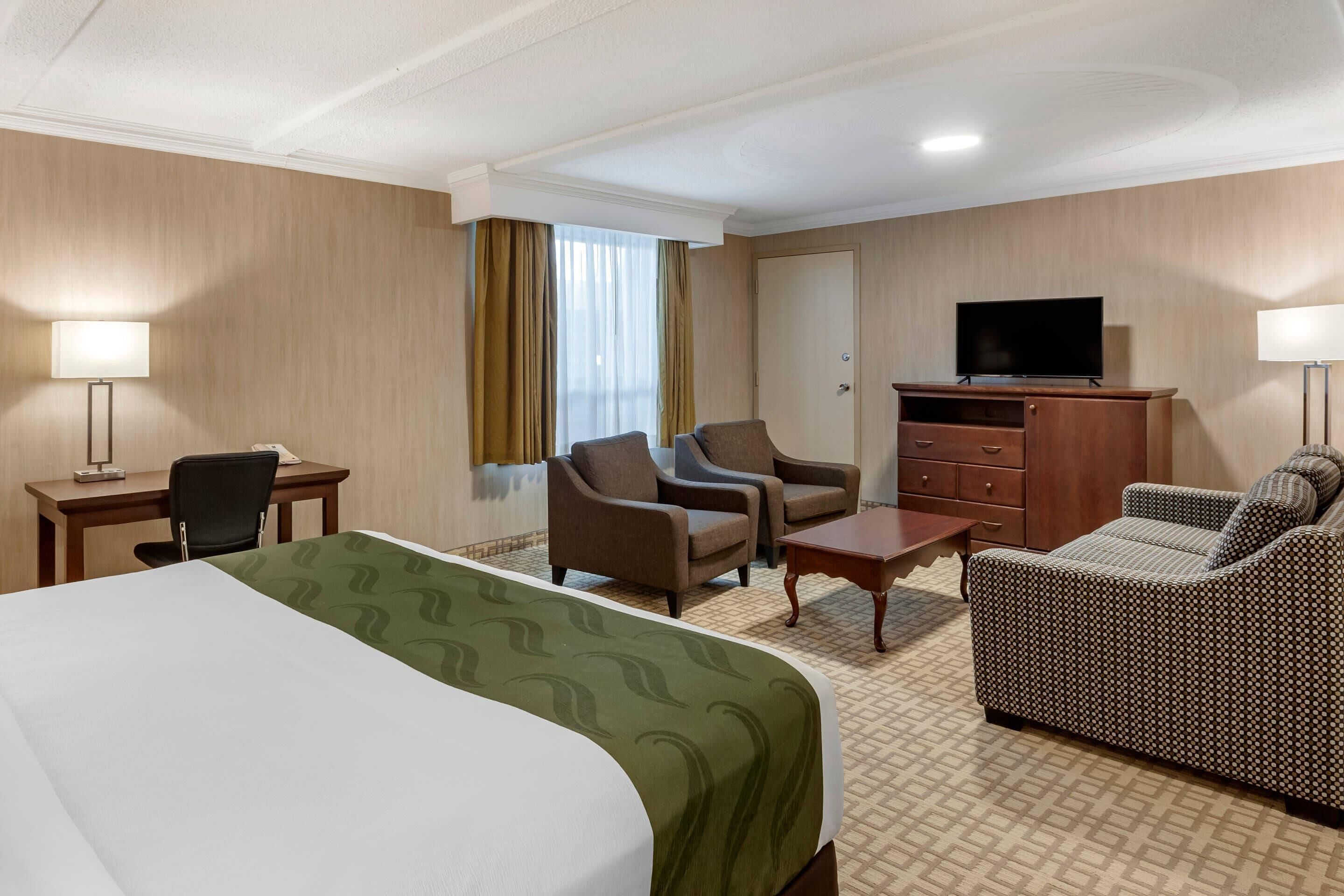 Une chambre d’hôtel avec un grand lit, deux fauteuils, un canapé, un bureau avec une lampe, un téléviseur posé sur un meuble et une fenêtre avec des rideaux.