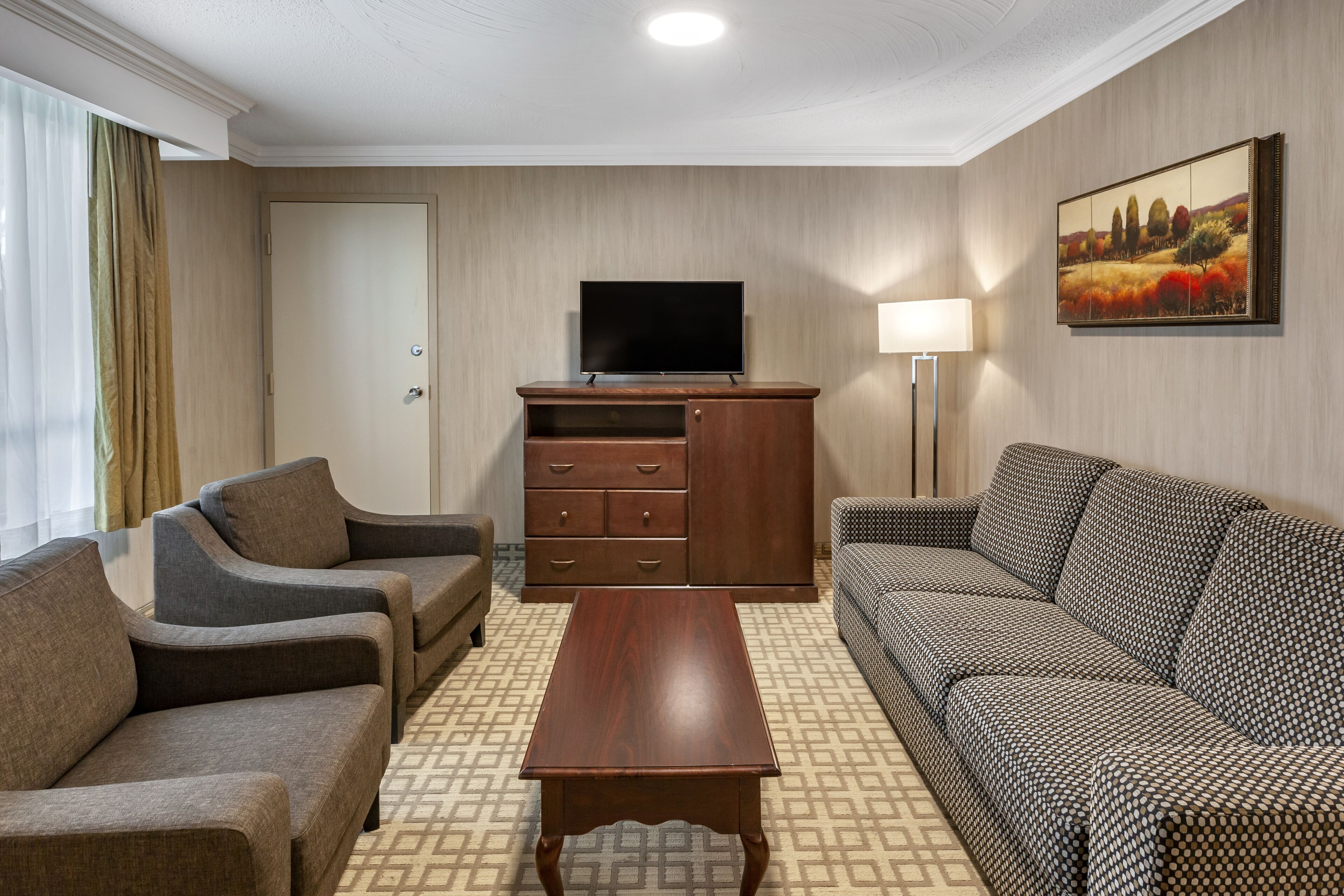A hotel room with a sofa, two armchairs, a wooden coffee table, a TV on a stand, a cabinet, a lamp, and a painting on the wall.