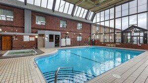 An indoor swimming pool with a tiled floor, a diving board, and a brick building with large windows in the background.