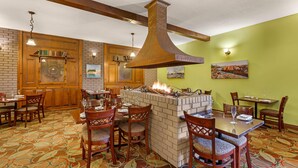 A restaurant with brick walls, wooden ceiling beams, and a central brick fireplace with a metal chimney. Tables and chairs are arranged for dining.