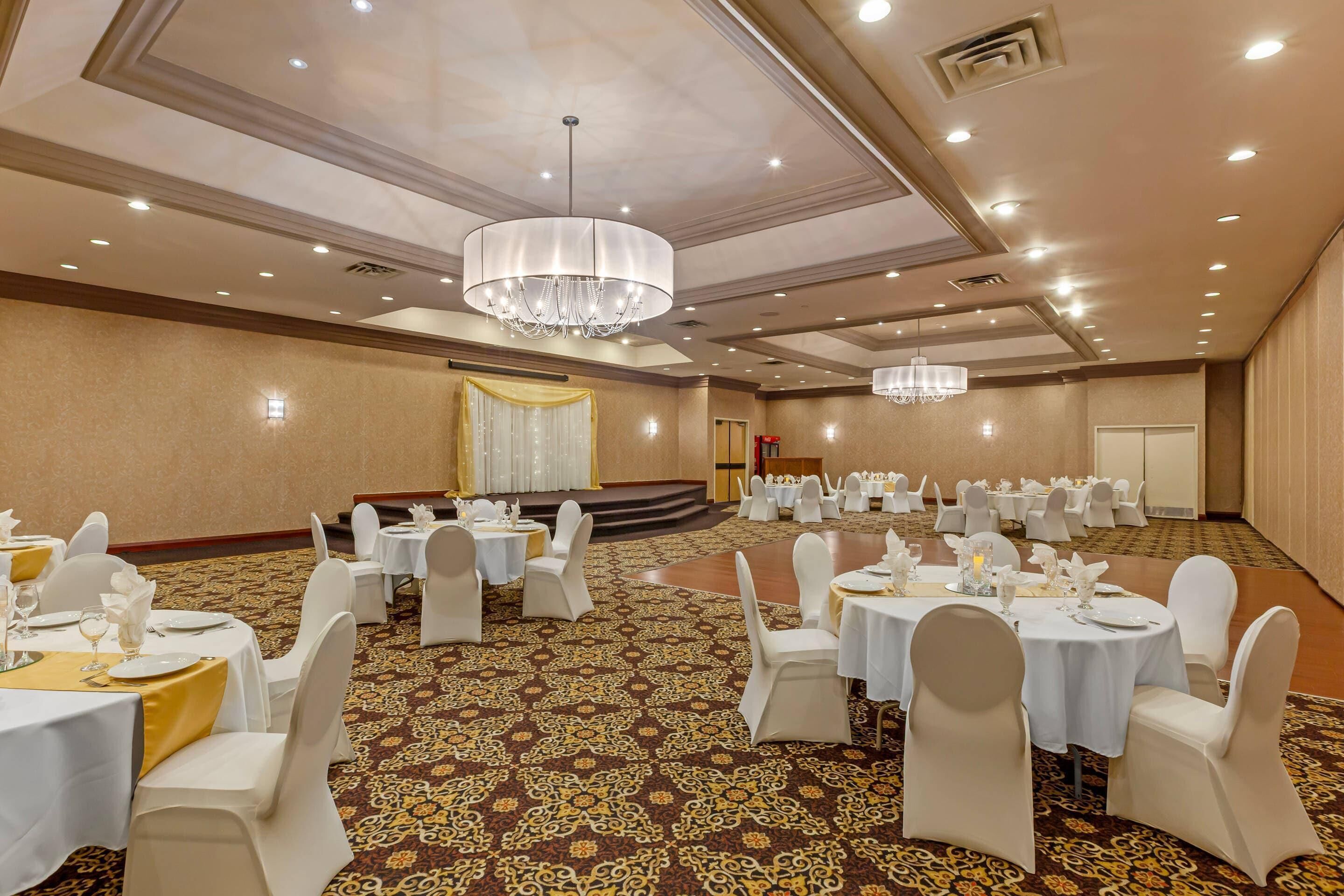 A banquet hall with round tables, white chairs, chandeliers, and a carpeted floor.