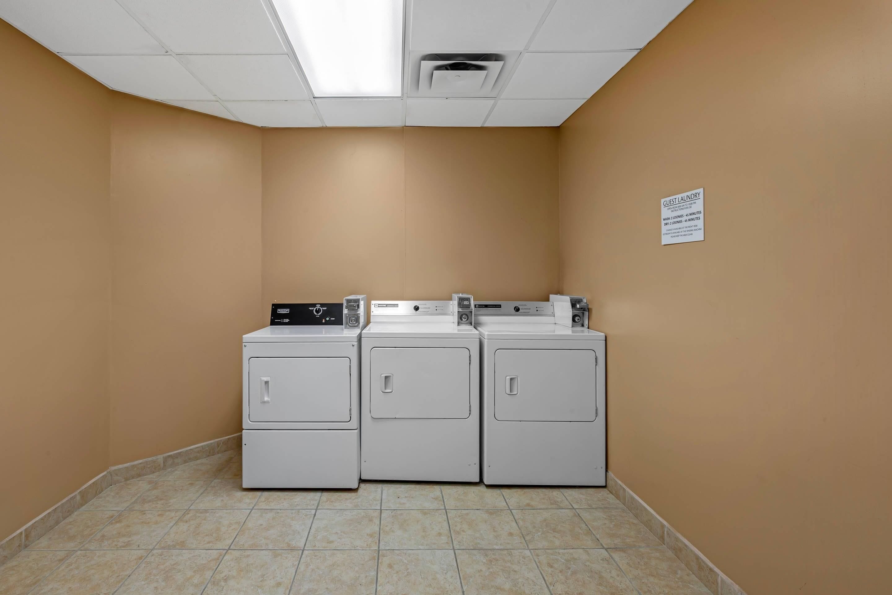 A laundry room with three stacked washers and dryers, a sign indicating guest laundry hours, and a tiled floor.