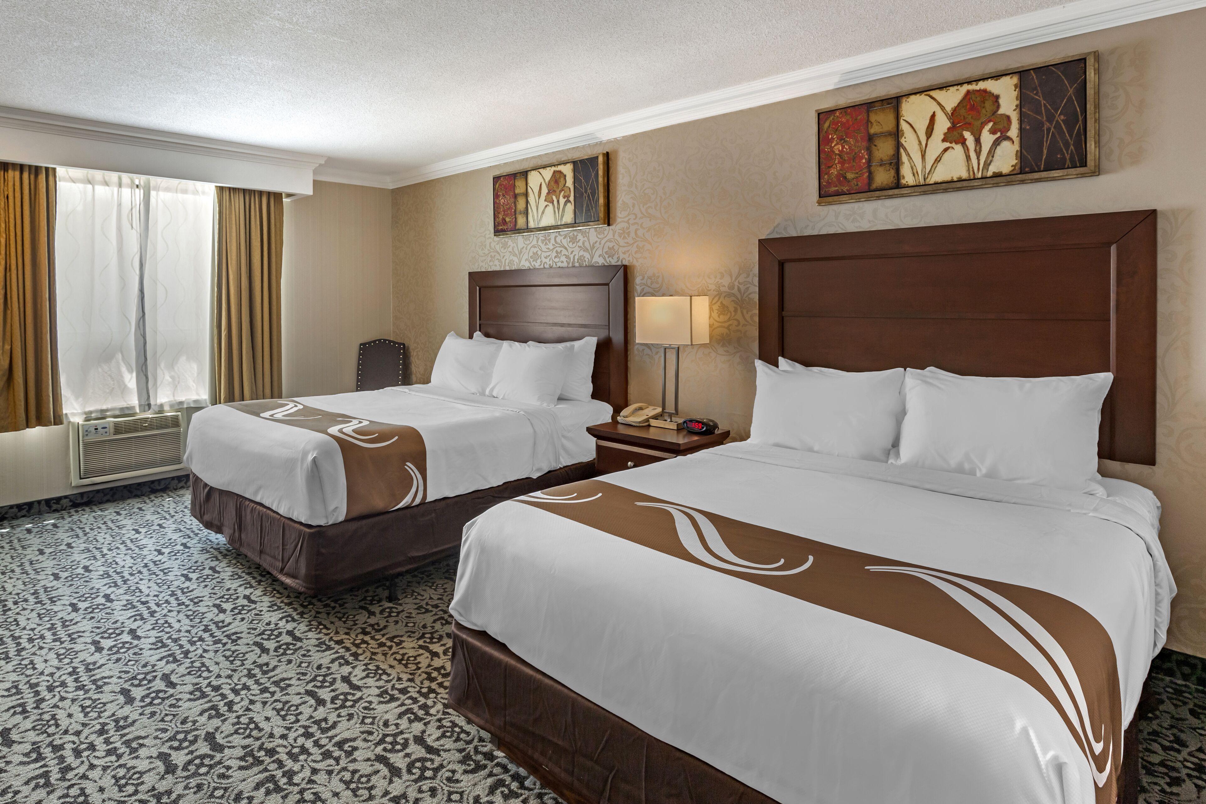 A hotel room with two beds, a desk, a chair, a lamp, and artwork on the walls.