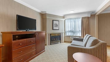 A hotel room with a wooden cabinet, a flat-screen TV, a patterned sofa, a small round table, a fireplace, and a framed picture on the wall.