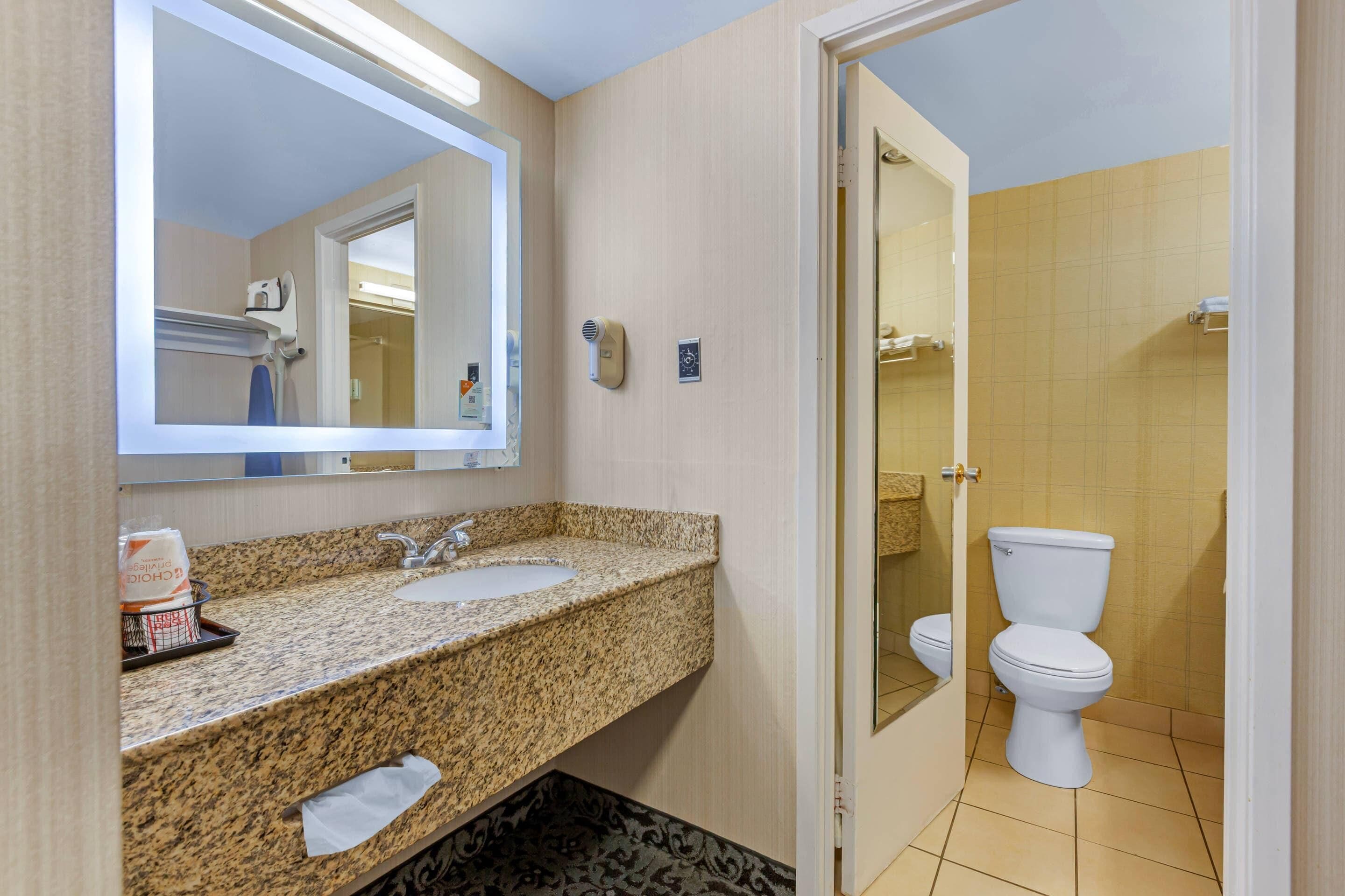 A bathroom with a granite countertop sink, a mirror, a handheld hair dryer, and a toilet.