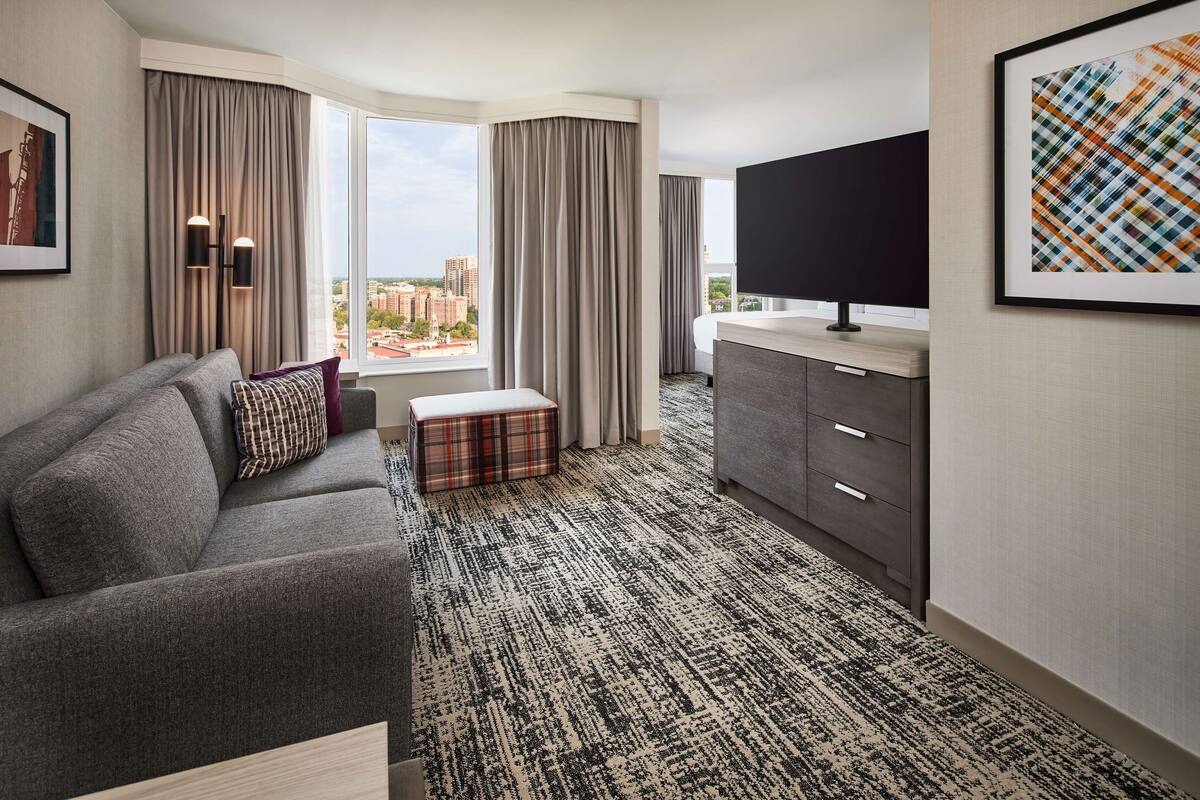 Ein Hotelzimmer mit grauem Sofa, Pouf mit Karomuster, Flachbildfernseher, grauem Schrank und einem großen Fenster mit Blick auf die Stadt.