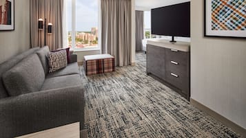 A hotel room with a grey sofa, a plaid ottoman, a flat-screen TV, a grey cabinet, and a large window offering a city view.