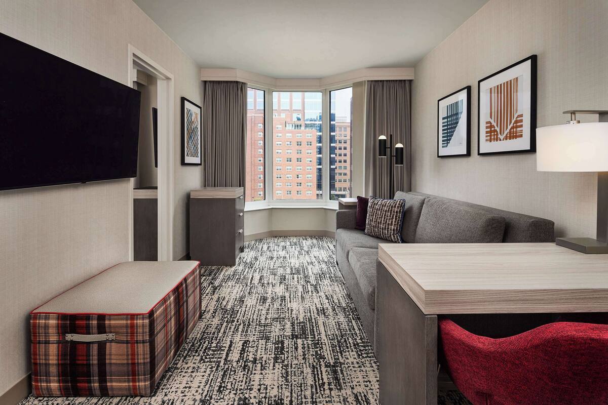 Ein Hotelzimmer mit großem Fenster, Flachbildfernseher, einer Couch, einem kleinen Tisch und einem roten Sessel.