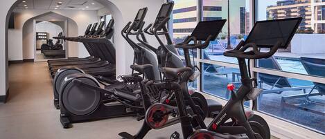 A gym with stationary bikes, a treadmill, and a view of the city through large windows.
