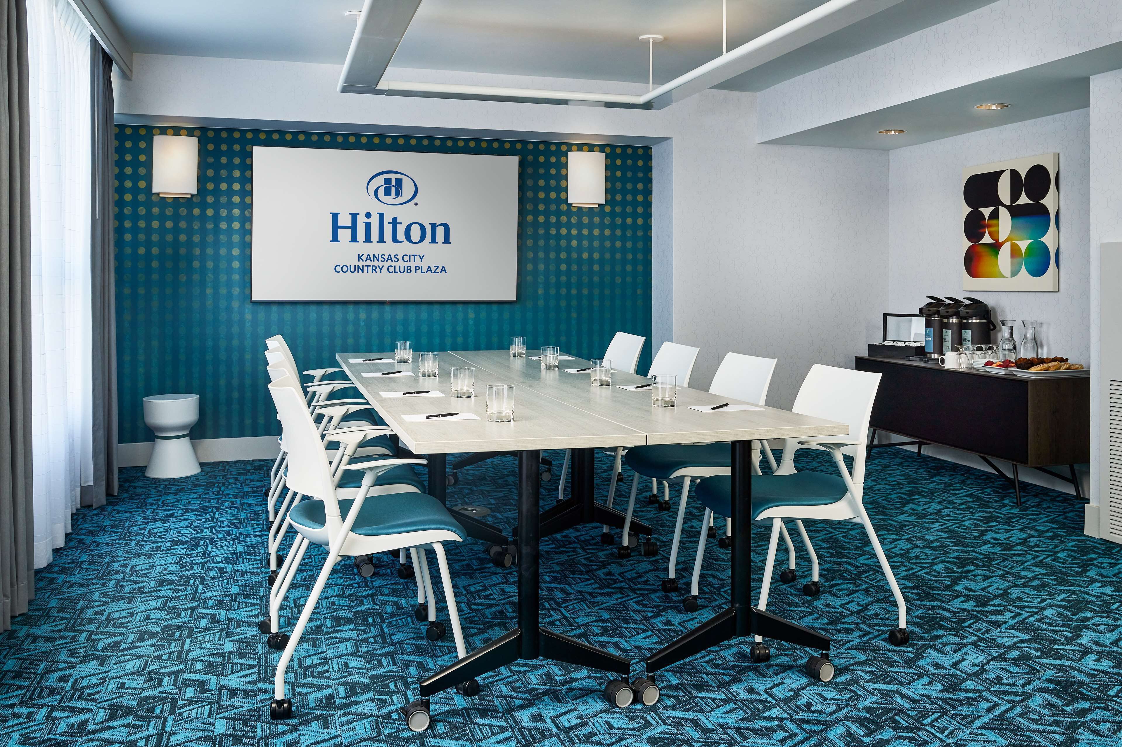 Una sala de conferències amb una taula llarga, cadires blanques i una catifa blava. Un gran pantalla mostra el logotip i el nom de Hilton.