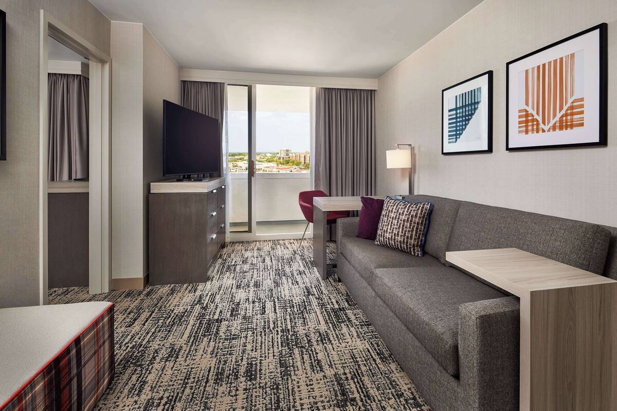 Ein modernes Hotelzimmer mit grauem Sofa, einem Flachbildfernseher, einem kleinen Tisch und einem Balkon mit Vorhängen.