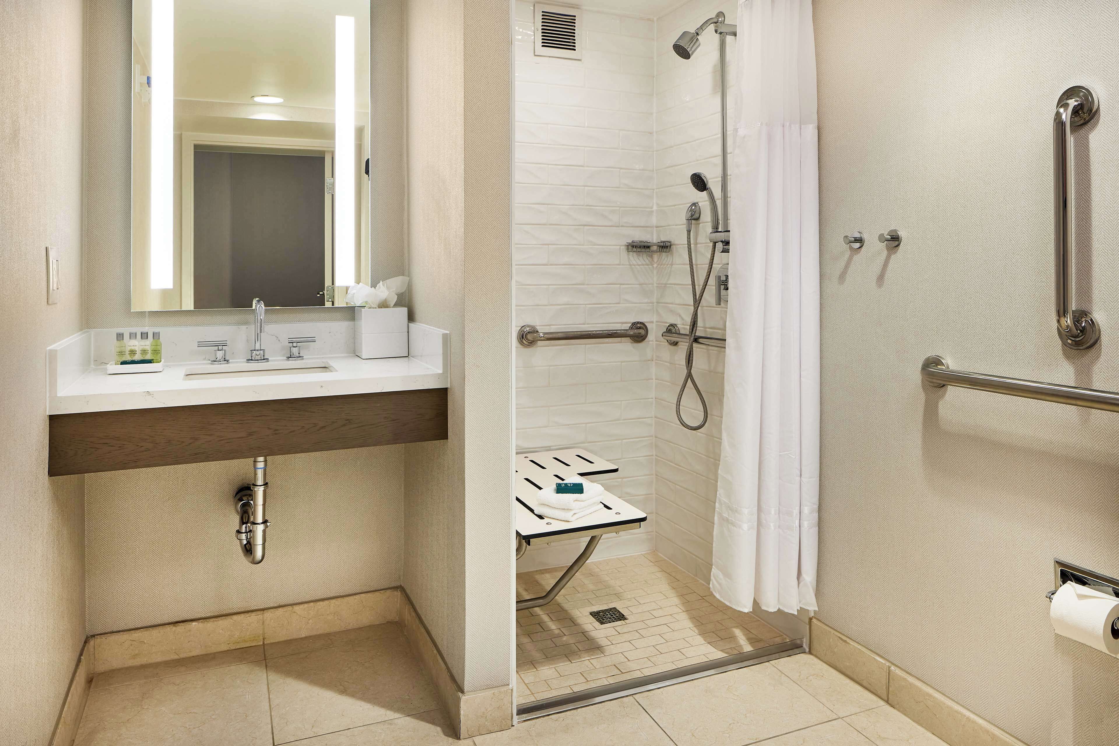 A modern bathroom with a shower, toilet, and sink. It includes a handrail, grab bars, and a towel rack.