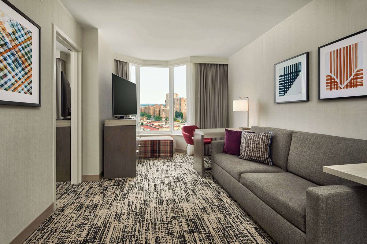 Ein Hotelzimmer mit großem Fenster, grauem Sofa, Flachbildfernseher, kleinem Tisch und rotem Sessel.