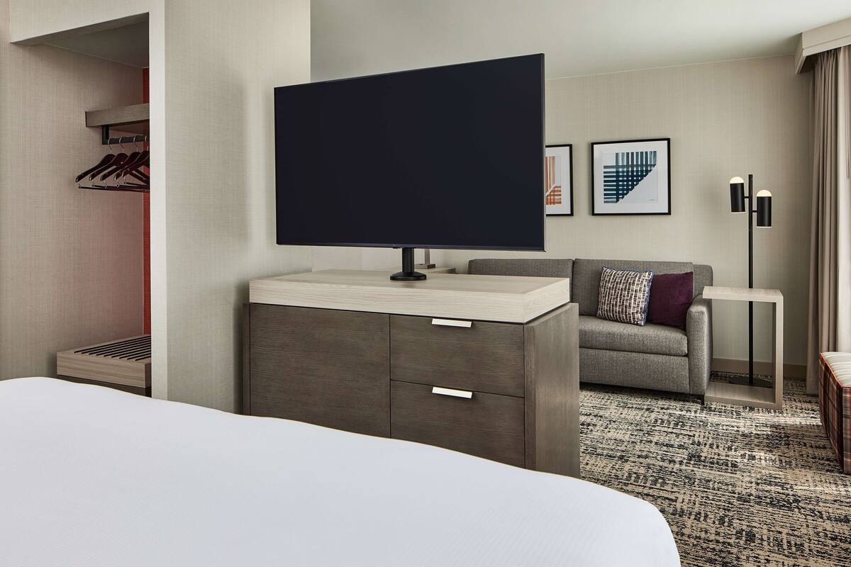 Ein modernes Hotelzimmer mit einem großen Bett, einem flachbildfernseher an einem Schrank, einem grauen Sofa mit einem gemusterten Kissen, einem kleinen Beistelltisch und einer Stehlampe.