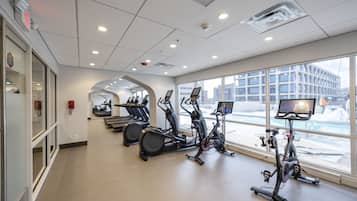 A gym with multiple exercise machines, large windows, and a view of the cityscape.