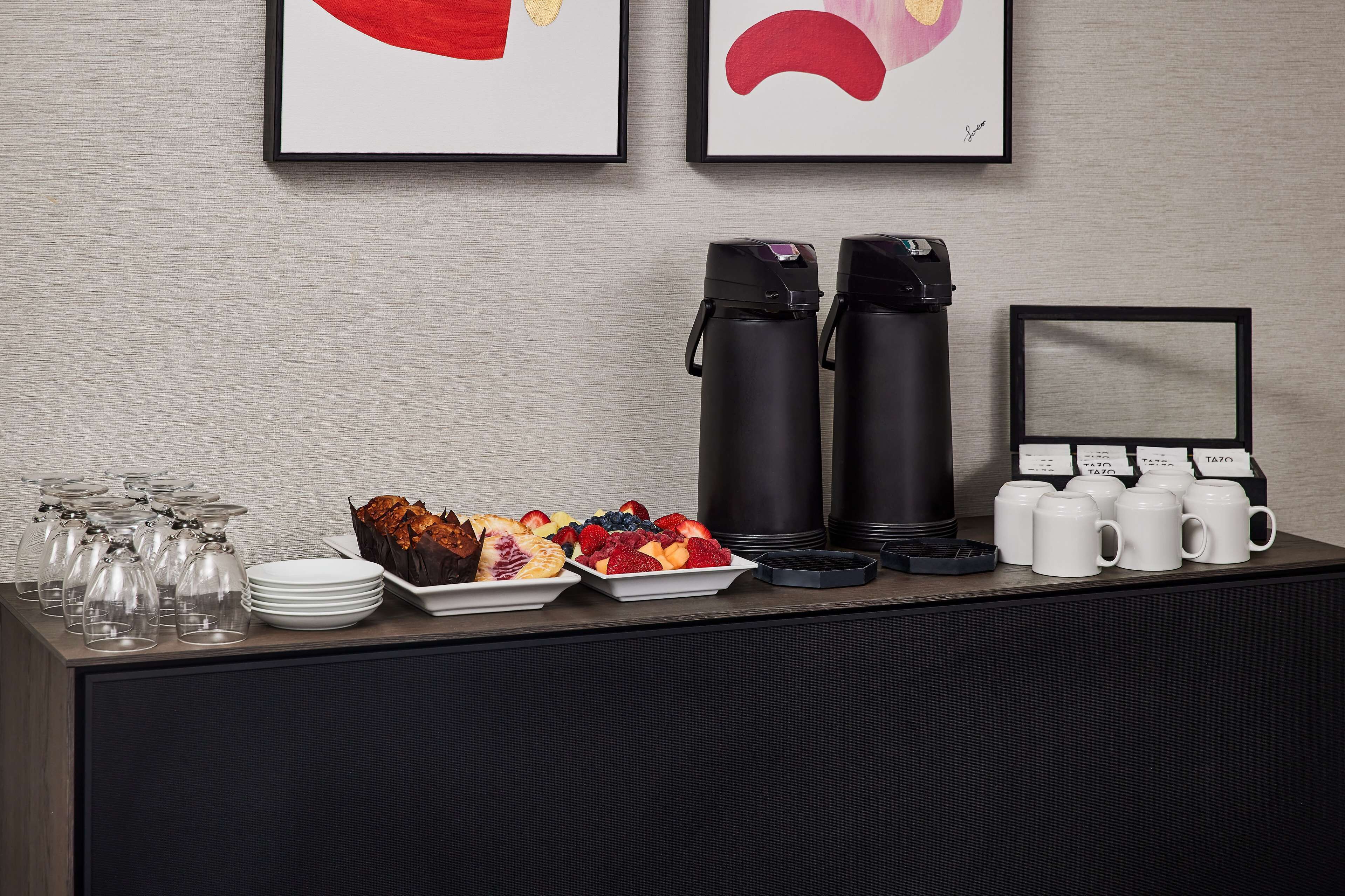 Montaje de un bufé de desayuno con máquinas de café, tazas y una variedad de frutas y pasteles.