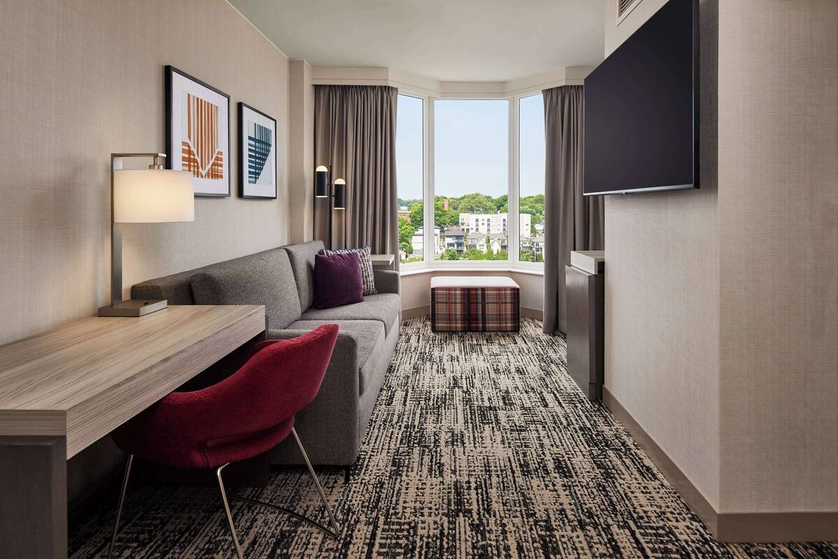 Ein Hotelzimmer mit einer Couch, einem roten Sessel, einem Schreibtisch, einem Flachbildfernseher und einem großen Fenster mit Blick auf die Stadt.