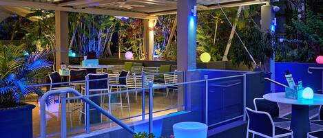 Un espace extérieur aménagé en terrasse, avec des sièges et des tables, entouré d’une végétation luxuriante et illuminé par un éclairage décoratif.