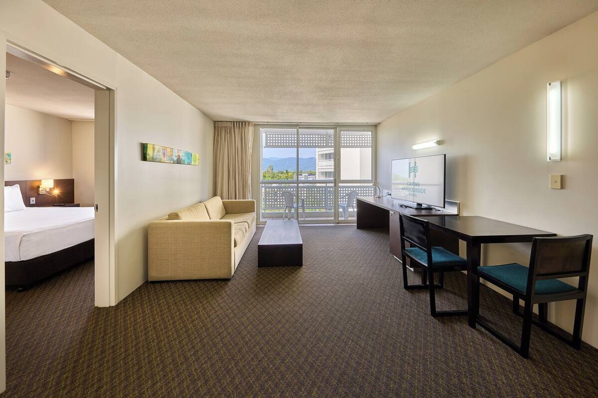 Habitación de hotel con cama, sofá, escritorio con sillas, televisión y balcón con vistas.
