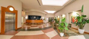 Welcome to our lobby, featuring a checkered floor, reception desk, and a large circular clock above the elevator, surrounded by lush greenery and natural light.