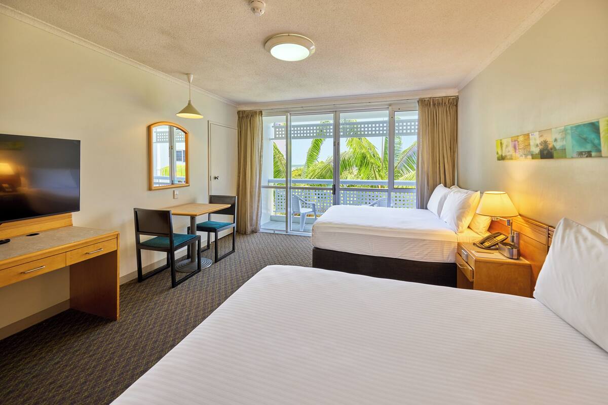 Habitación de hotel con una cama grande, un escritorio con silla, televisión y balcón con vista a palmeras.