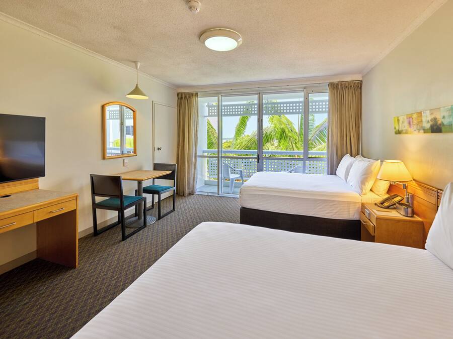 A hotel room with a large bed, a desk with a chair, a television, a window with a view of palm trees, and a balcony.