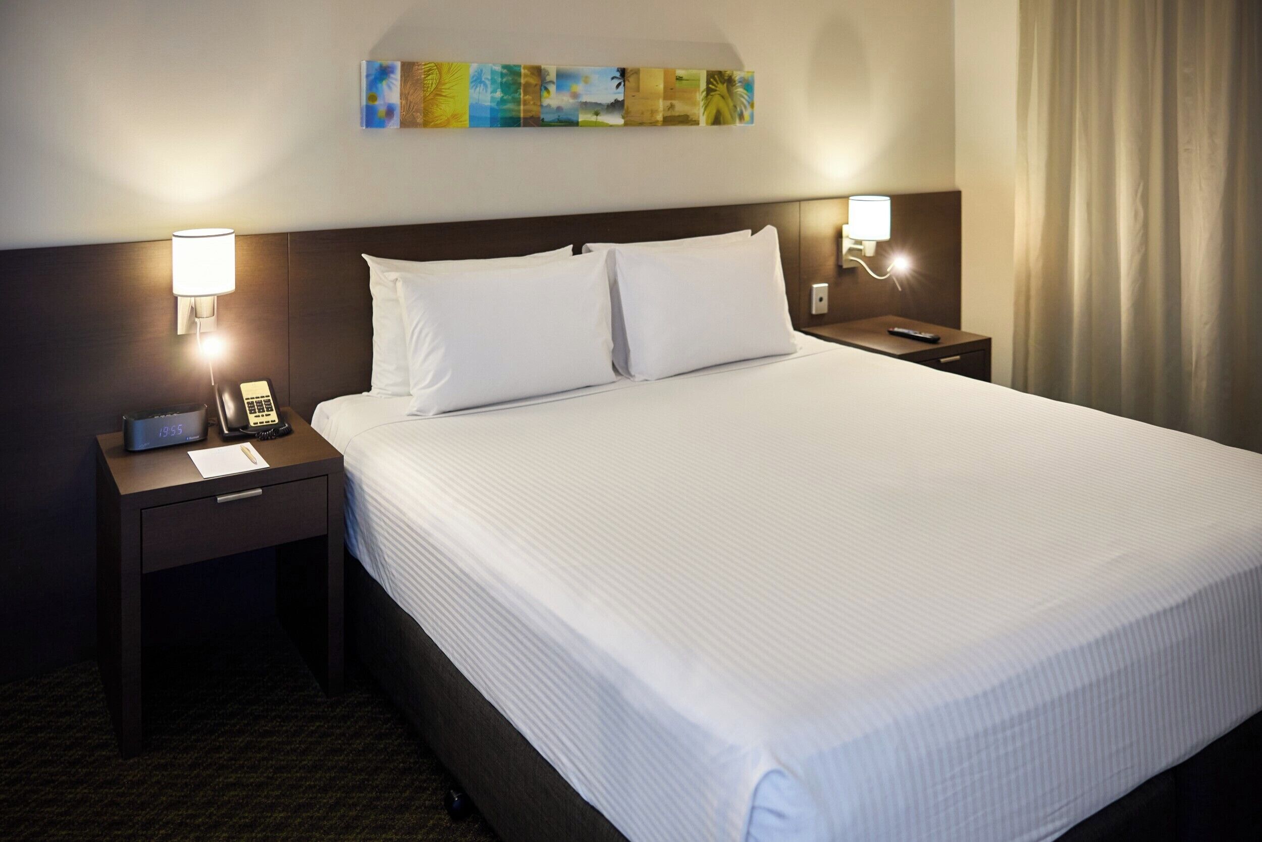 Een hotelkamer met een groot bed, twee nachtkastjes, een telefoon en een kunstwerk aan de muur.
