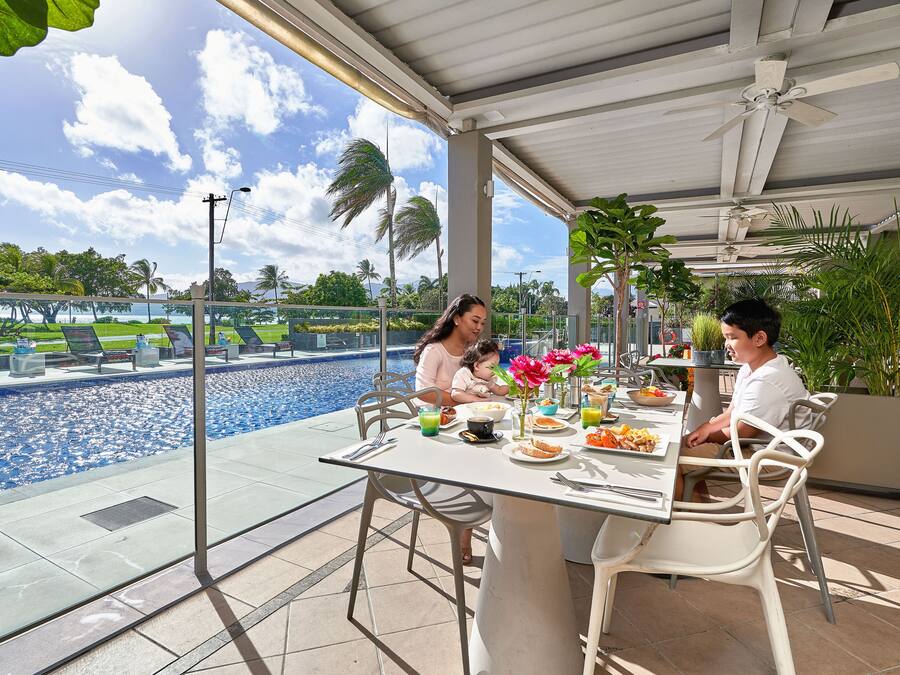Un área de comedor al aire libre con vistas a una piscina, rodeada de palmeras y un cielo despejado.