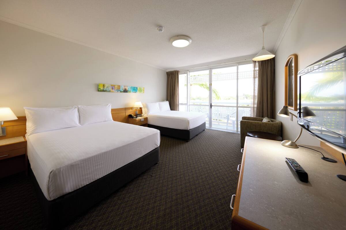 Habitación de hotel con dos camas, un escritorio con computadora, televisor y ventana con vista a palmeras.