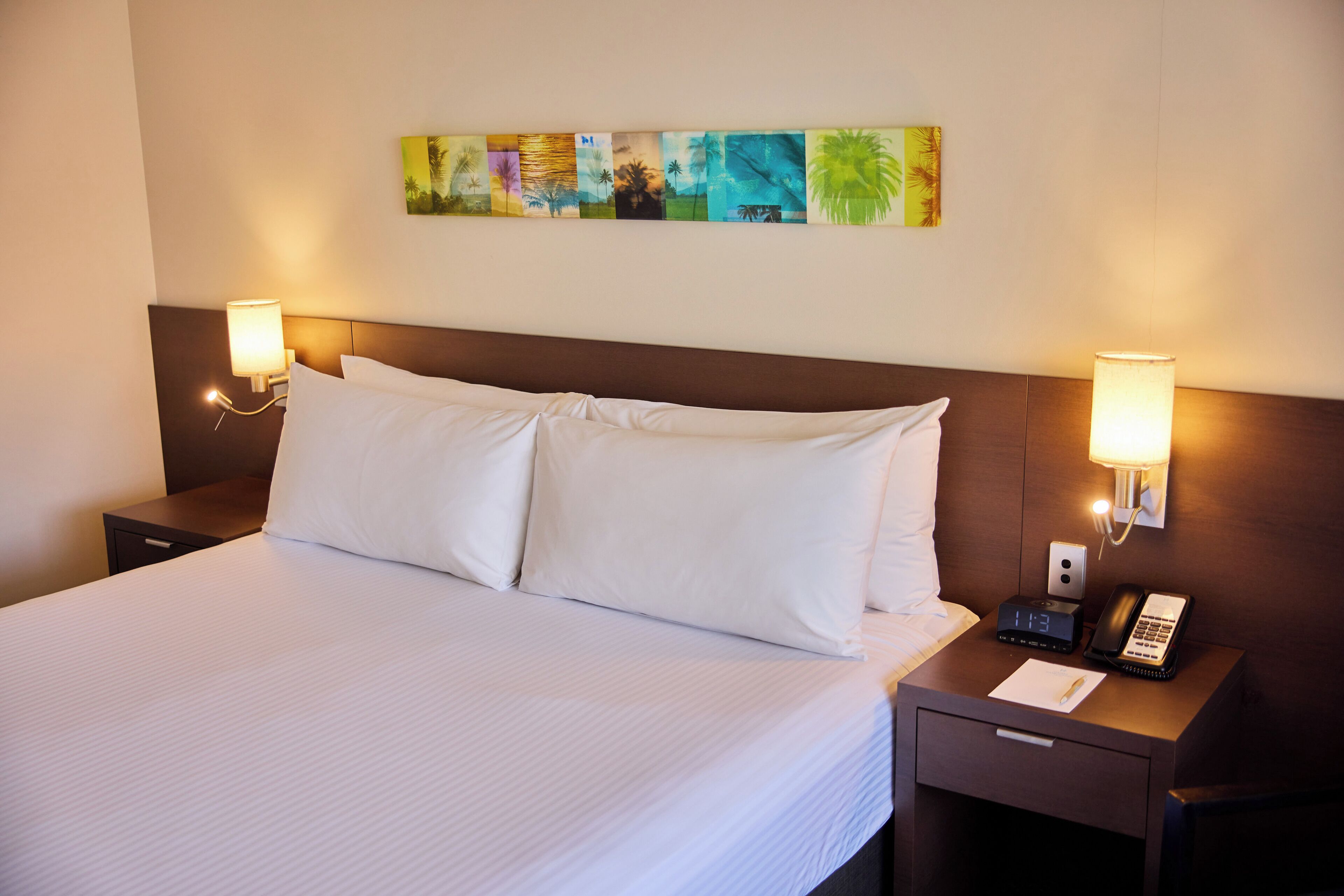 A neatly made bed with white linens, a nightstand with a lamp, a clock, and a phone.