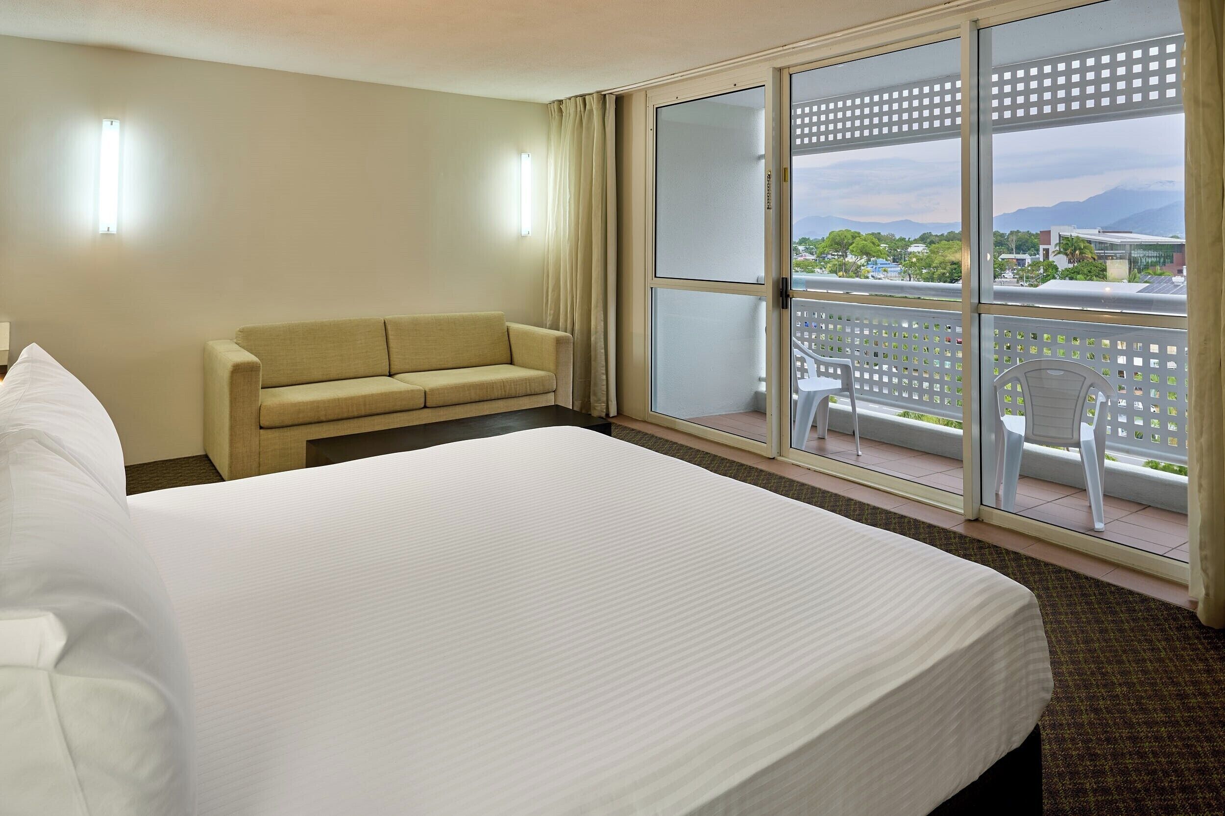 Habitación de hotel con una cama grande, un sofá, un balcón con vistas a la ciudad y las montañas, y una mesa con sillas.