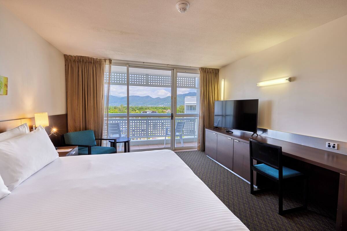 Habitación de hotel con una cama grande, un escritorio con silla, televisor y balcón con vista a montañas.
