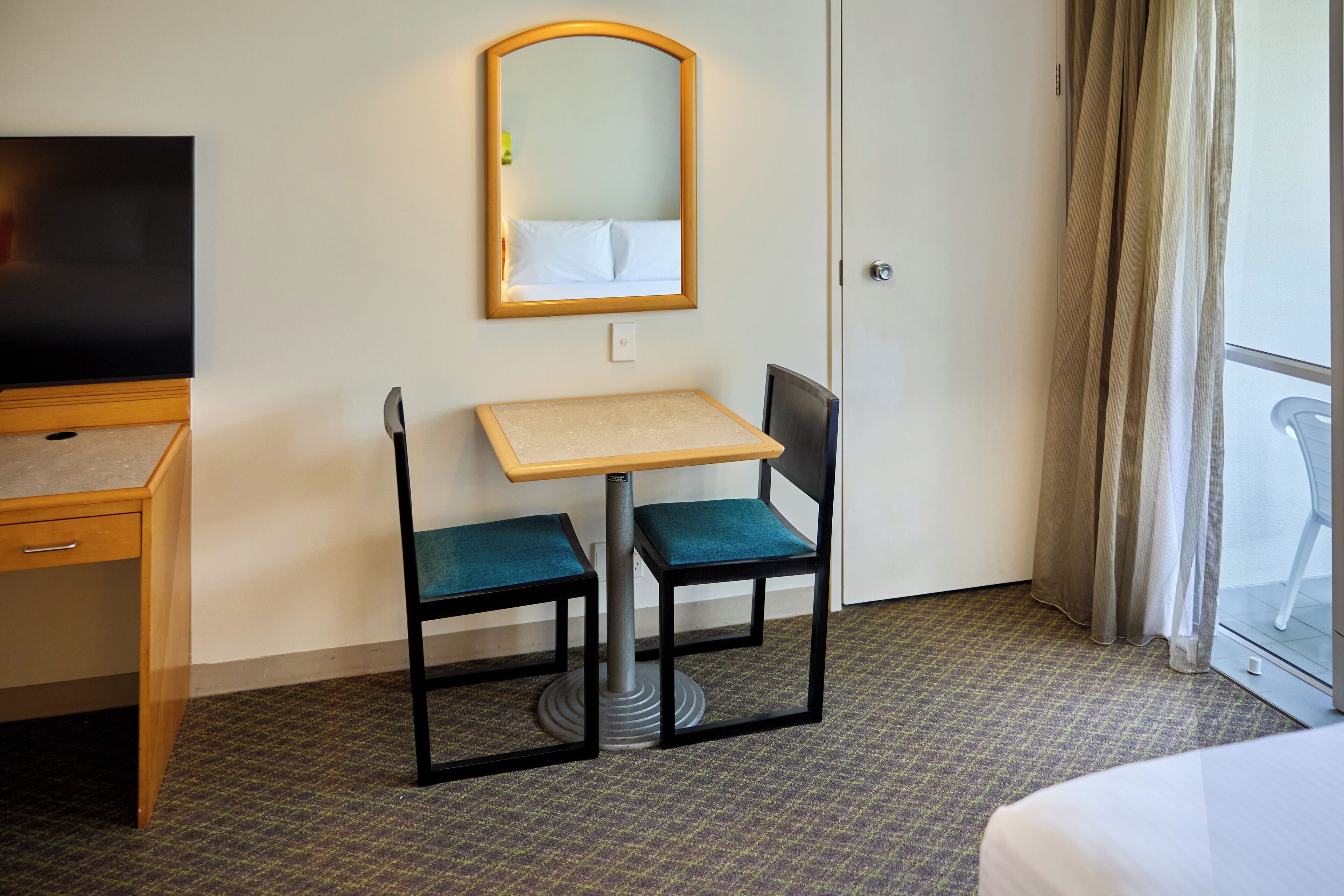A hotel room with a bed, a desk with a chair, a television, a small table, and a mirror.