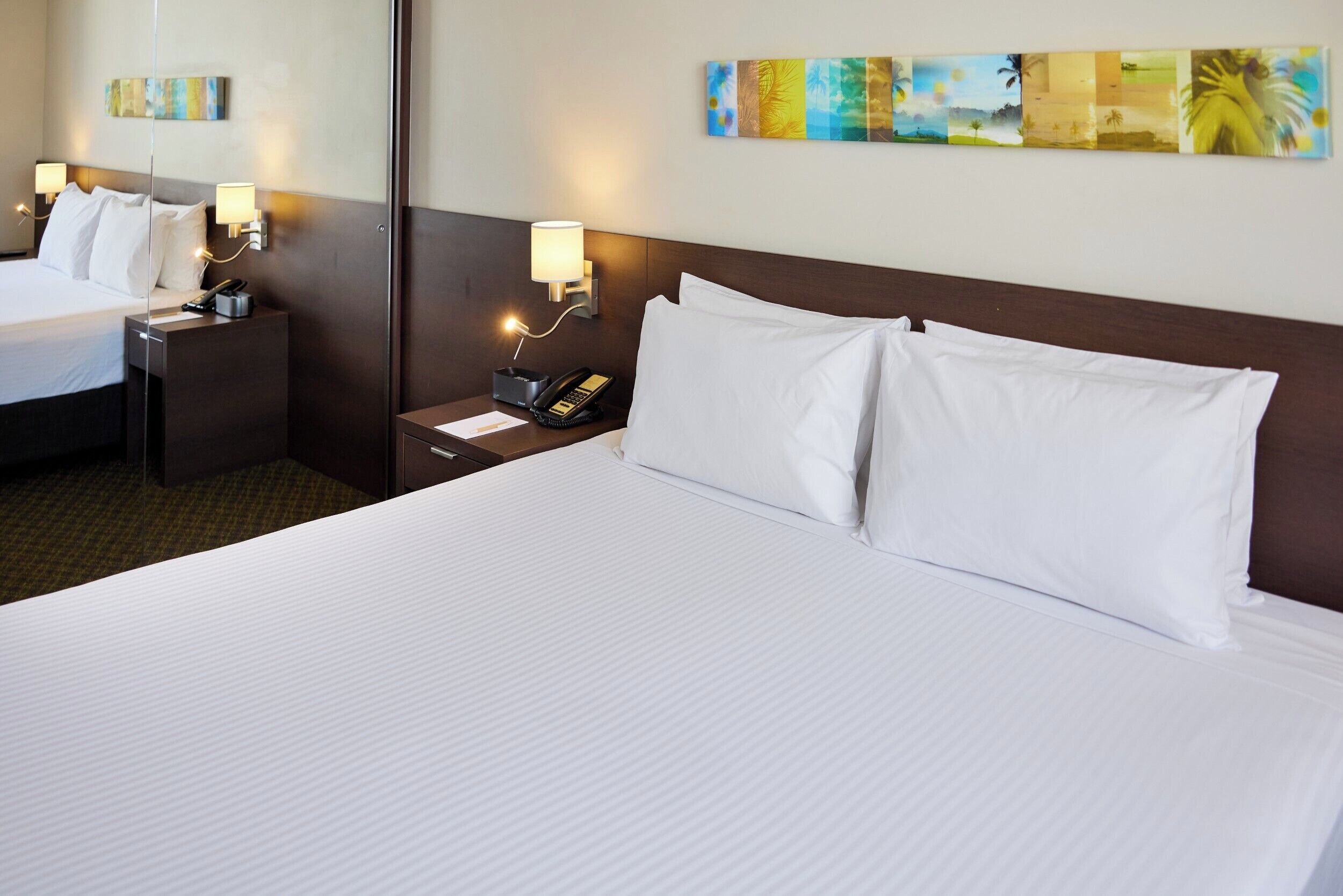 Habitación de hotel con una cama grande, dos mesitas de noche con lámparas, un teléfono y un cuadro con temática tropical en la pared.